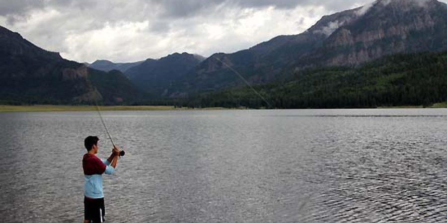 Man wading in lake fishing, with cloud-wrapped mountains in the distance.